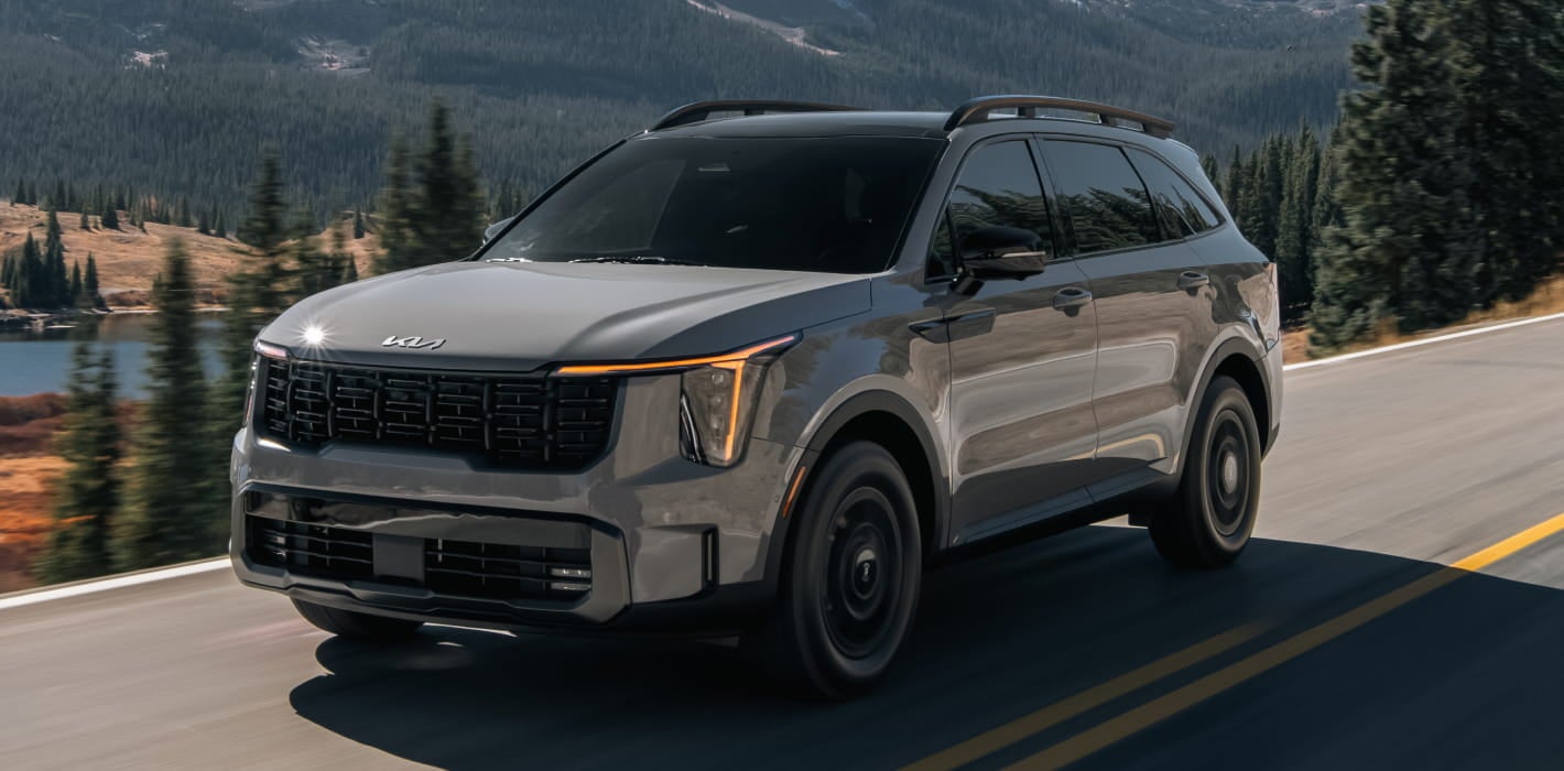 2026 Kia Sorento drives on a scenic road flanked by tall pine trees and a mountain range in the background.