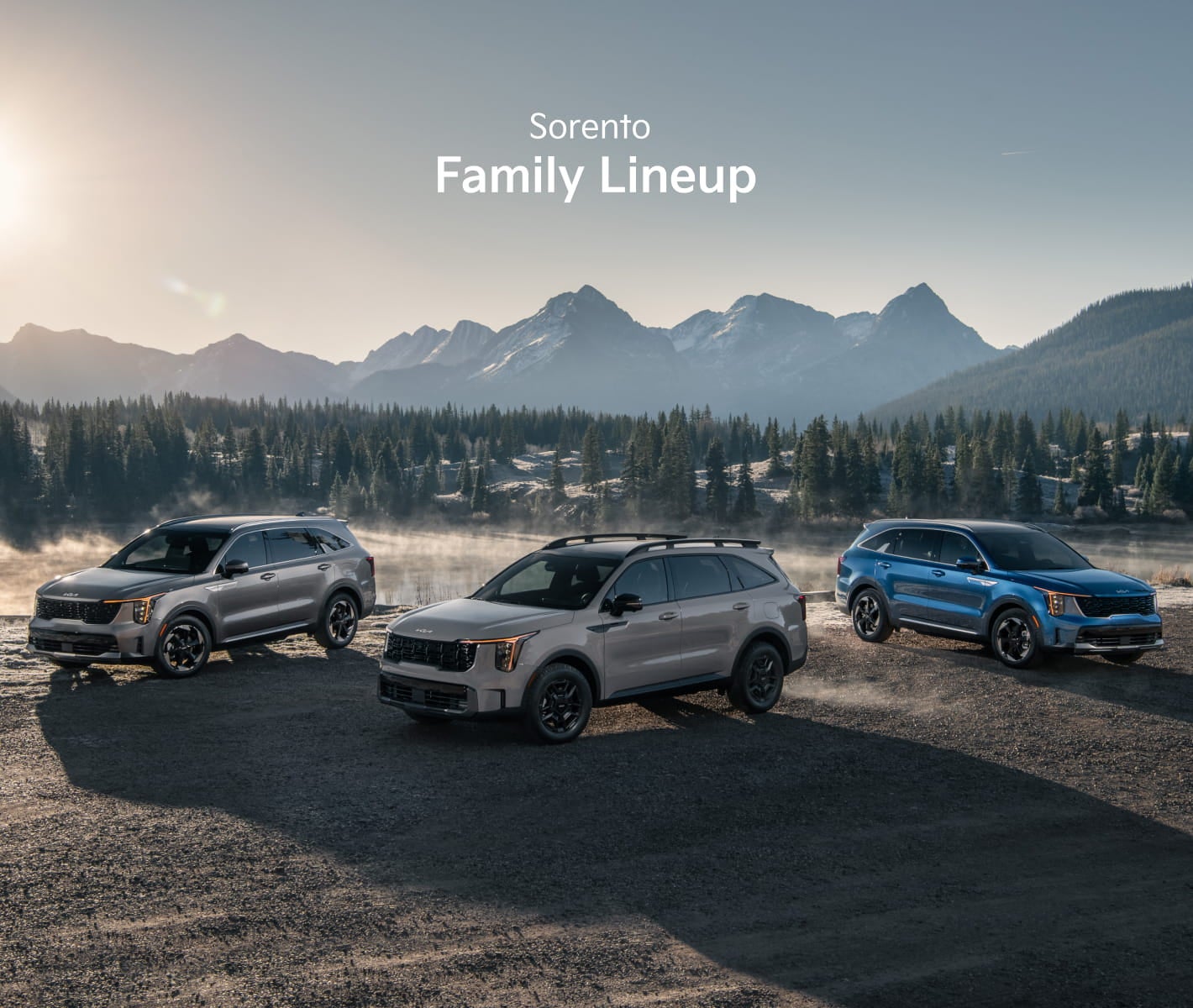 Three SUVs from the Sorento lineup: 2026 Sorento Hybrid, 2026 Sorento and 2026 Sorento PHEV are parked on a gravel area near a misty lake, with a mountain range in the background under a clear sky.