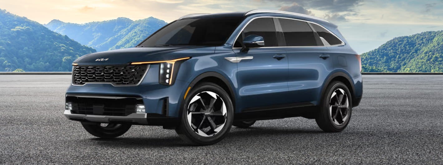 A sleek, blue 2026 Kia Sorento Hybrid with modern design and black accents is parked on a paved area against a backdrop of green, rolling mountains under a partly cloudy sky.